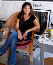 玛丽Dondero sitting on a rattan chair in her studio with a framed picture on the floor behind her and a table with paint cans and brushes on it next to her