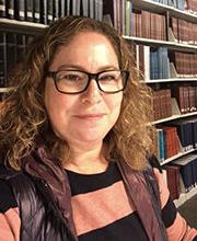 headshot photo of Linda Meiberg in front of library bookshelves with light brown medium length curly hair wearing black rim glasses and a black and salmon top under a black vest