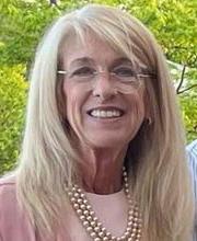 Dr. Kimberly Merrick smiling with long grey-blonde hair wearing rimless round glasses a pink top and pink pearls