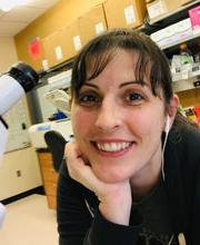 Dr. Joslyn Mills next to microscope in lab smiling with long dark brown hair pulled back in a pony tail with bangs and wearing earphones and a 黑色的 long sleeve top