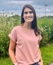 Dr. Kerri Olore standing in front of outdoor flowers and greenery smiling with long straight dark brown hair and wearing a short sleeve pink blouse and jeans