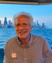 Leonard Paolillo on a boat smiling with short gray hair wearing a black and white checked button down shirt with water and a city skyline in the background