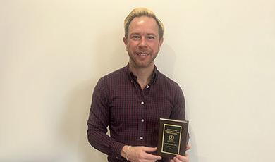 Adam Brieske-Ulenski stands in front of a wall holding his award plaque.