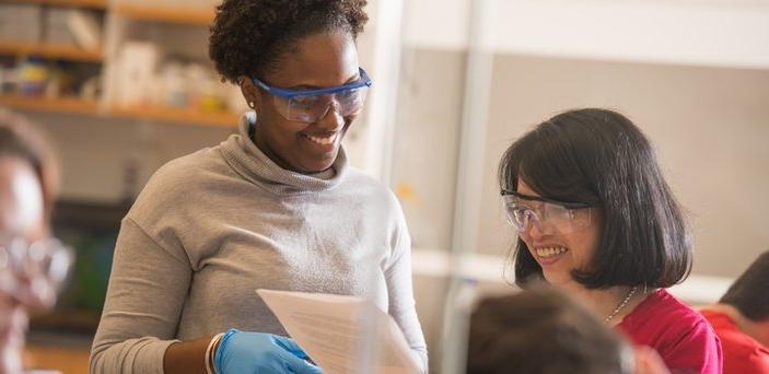 A student and professor, wearing rubber gloves and safety glasses, smiling and reviewing classwork.