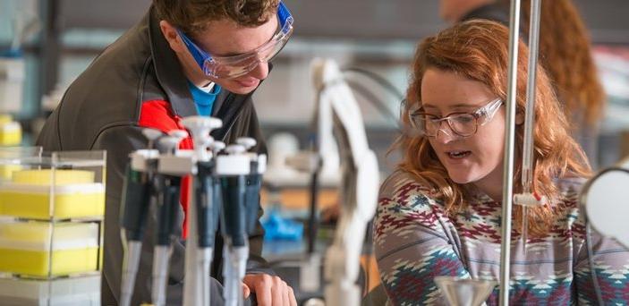 Two students working at a table in a chemistry lab wearing safety glasses with equipment in front of them.