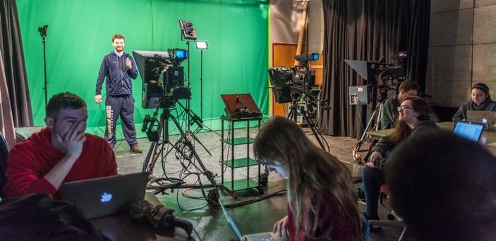 Foreground of students at tables working on laptops. Background of a student standing with a microphone in front of a green screen with tv cameras in front of him. 