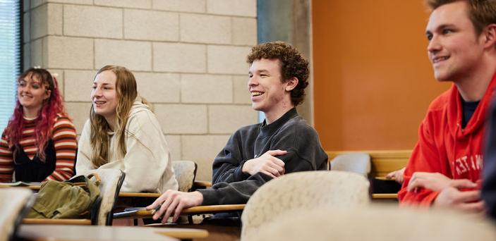 4 students sit in a row of individual desks smiling at the teacher