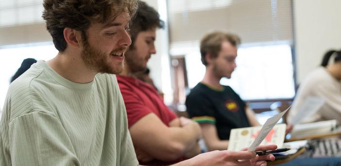 Students sit in a row of individual desks while the student closest to the camera reads from a paper with pen in hand