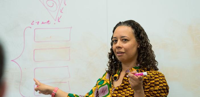 Dr. Allyson Ferrante teaches in front of a white board that she is pointing to while holding a marker in her other hand