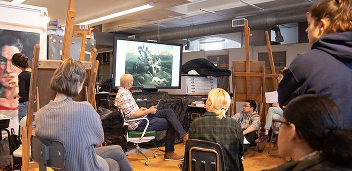 a professor 和 small group of students view paintings on a large screen while a student outside the group paints a large portrait