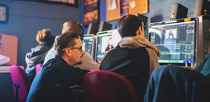 a professor helps a student in the BSU 沟通 Studies Editing Lab where a row of students sit at computers editing video